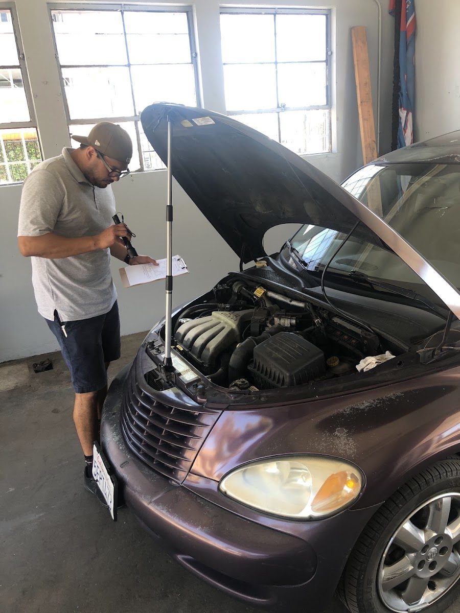 An Easy Breezy Smog technician with a clipboard checking under the hood of a PT Cruiser